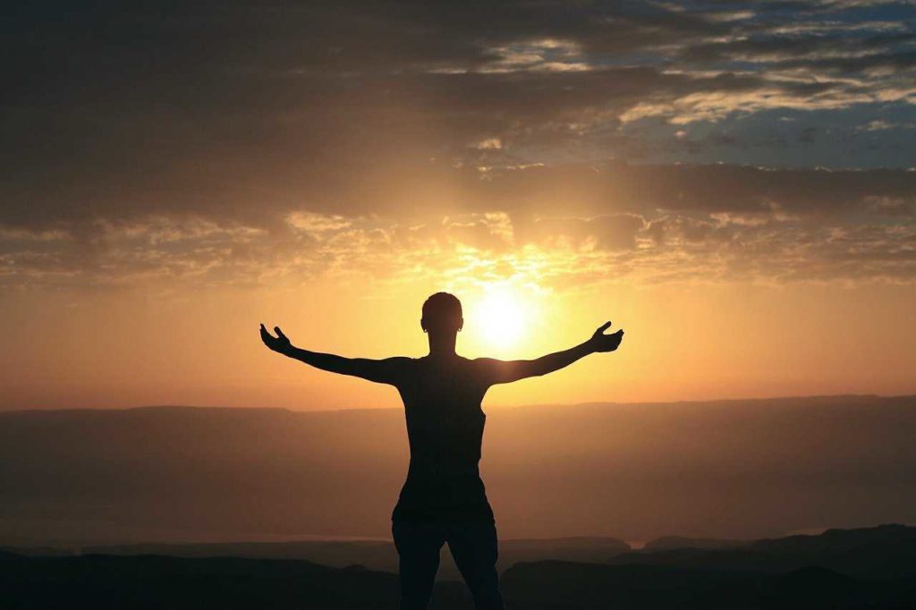 Das Foto zeigt einen Sonnenuntergang, vor dem eine Person mit ausgestreckten Armen steht. Die Person ist nur als Silhouette zu erkennen.